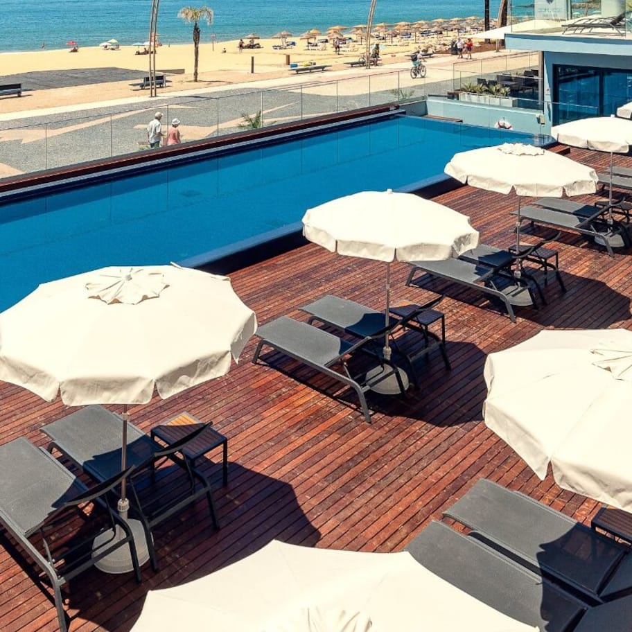 A rooftop pool area with lounge chairs and umbrellas, overlooking a beach and ocean.