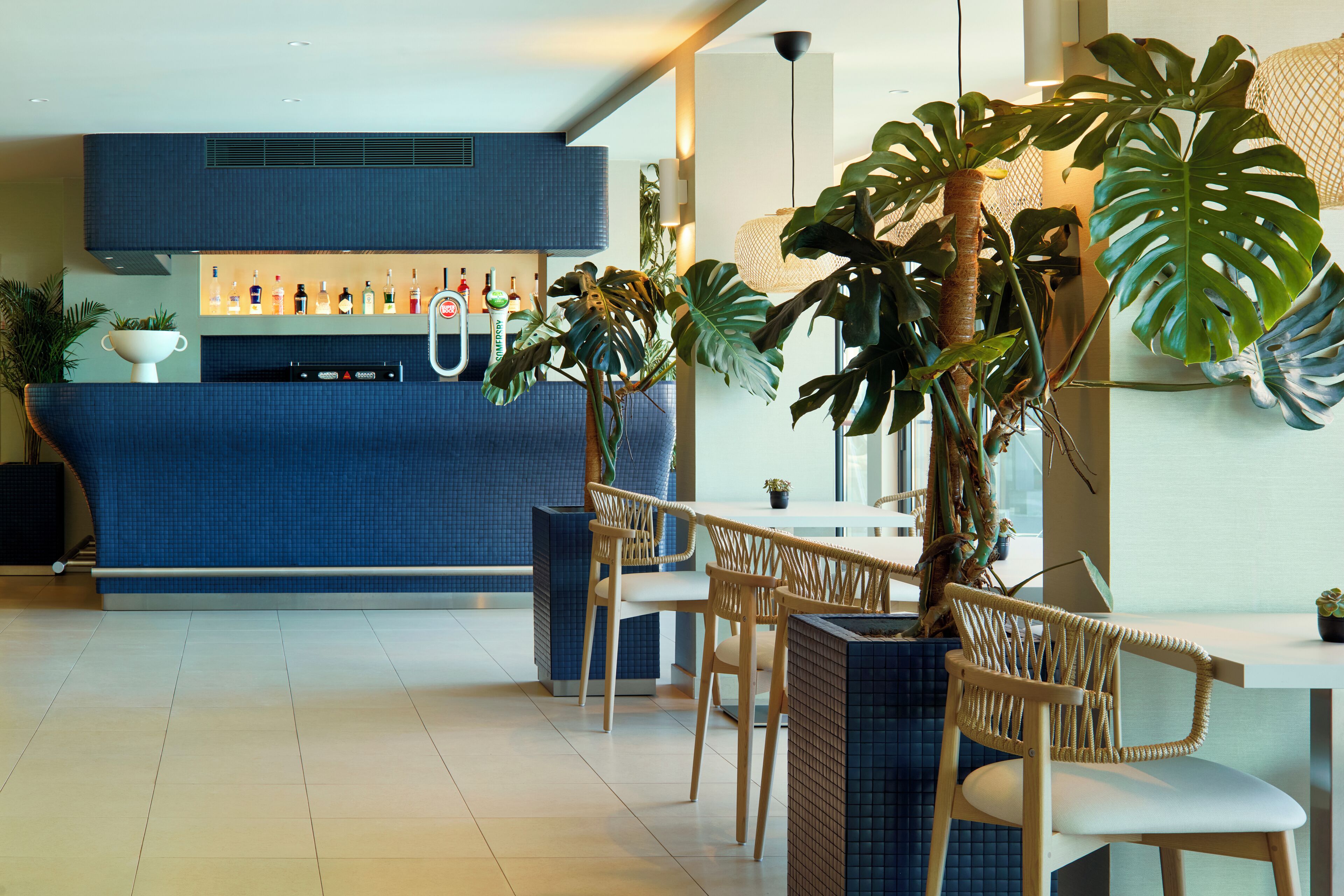 Uma área de bar moderna com bancada azul, cadeiras de madeira e grandes plantas tropicais.