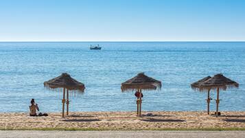 A beach with three thatched umbrellas, a person sitting on the sand, and a boat in the water.