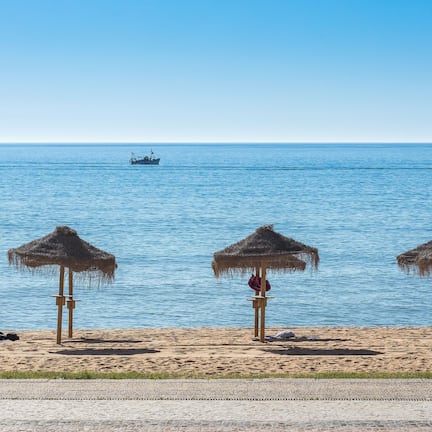 A beach with three thatched umbrellas, a person sitting on the sand, and a boat in the water.