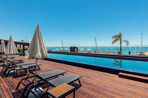 Un espace aménagé au bord de la piscine, avec des chaises longues et des parasols, une terrasse en bois et une vue sur la plage et l’océan.