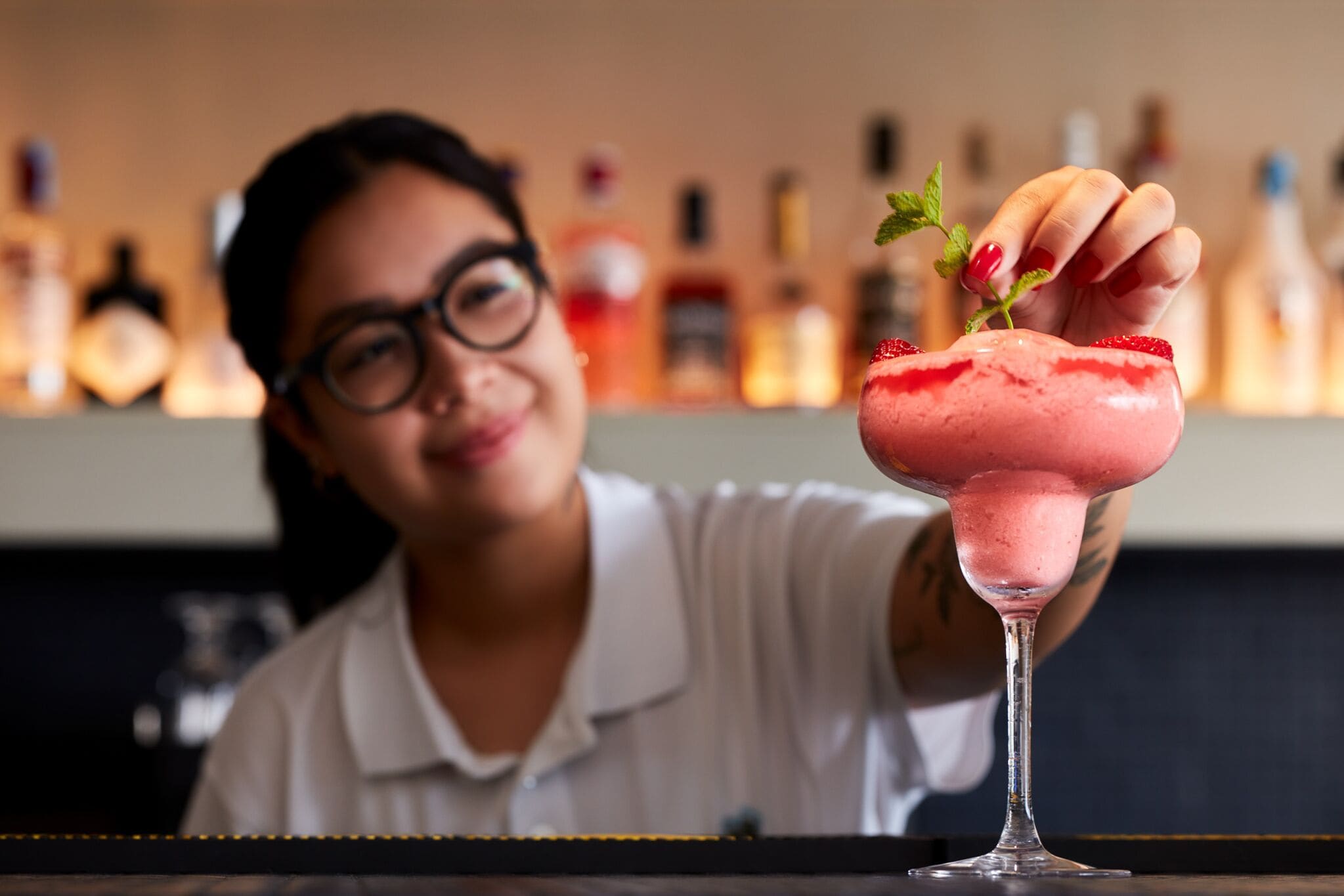A person holding a pink drink garnished with a mint leaf and a strawberry, with a blurred background of bottles.