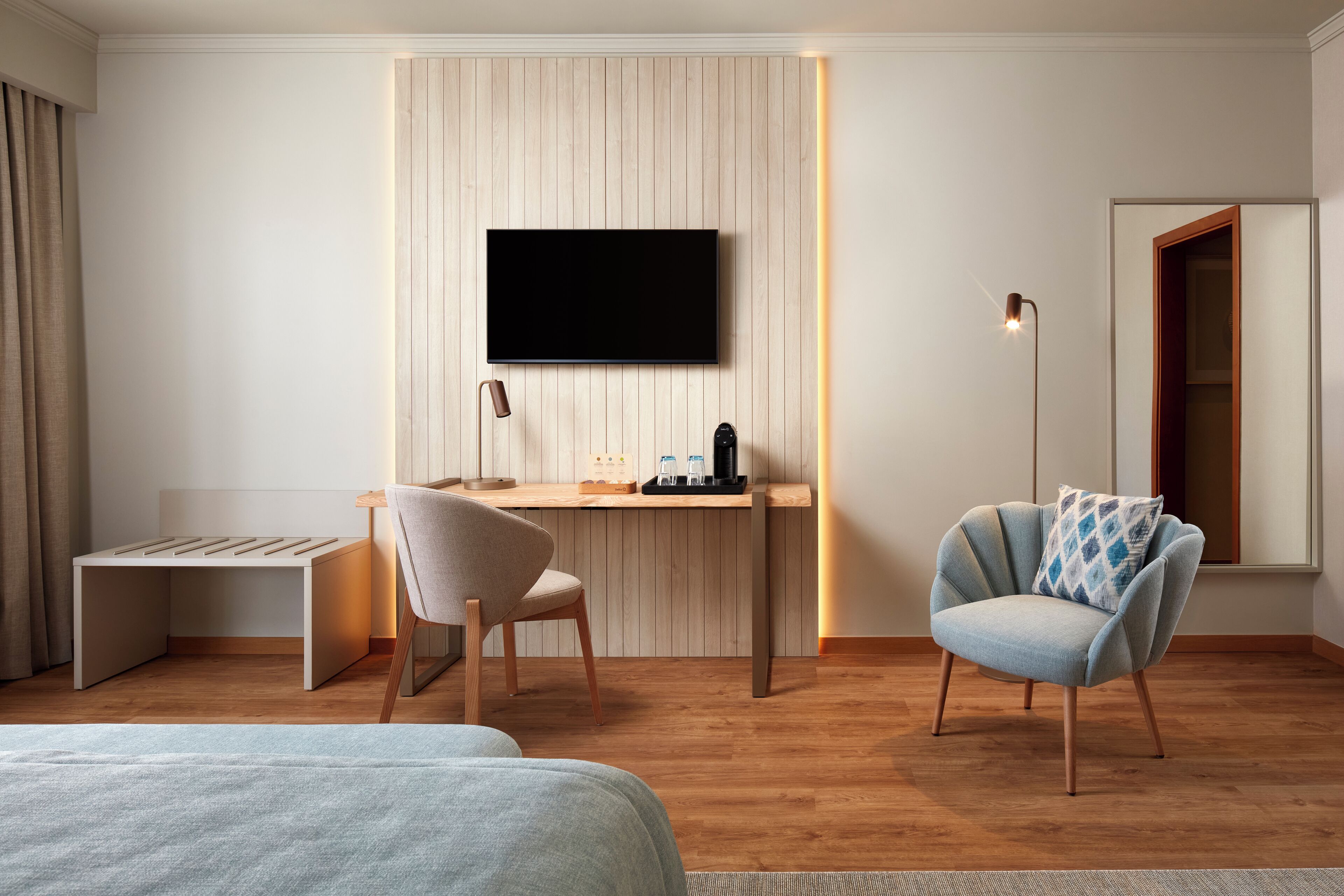 Ett modernt hotellrum med trägolv, en säng, ett skrivbord med en stol, en tv som är monterad på väggen och en golvlampa.