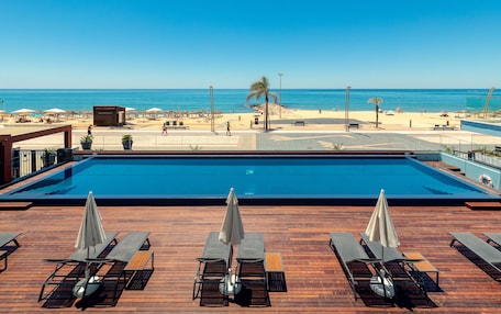 A pool area with lounge chairs and umbrellas, overlooking a beach with umbrellas and a palm tree.