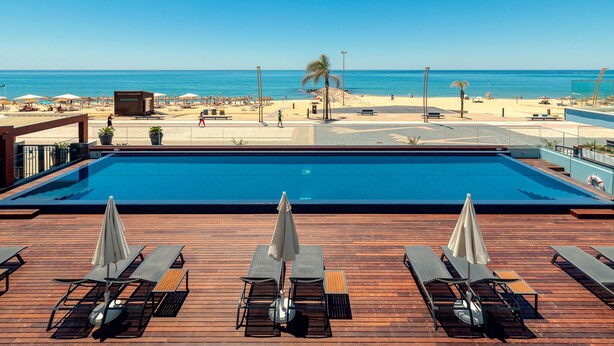 A pool area with lounge chairs and umbrellas, overlooking a beach with umbrellas and a palm tree. - Dom Jose Beach Hotel (Loulé)