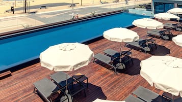 Uma área de piscina no terraço com espreguiçadeiras e guarda-sóis, um deck de madeira e vista para a praia.