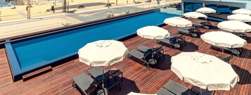 Uma área de piscina no terraço com espreguiçadeiras e guarda-sóis, um deck de madeira e vista para a praia.
