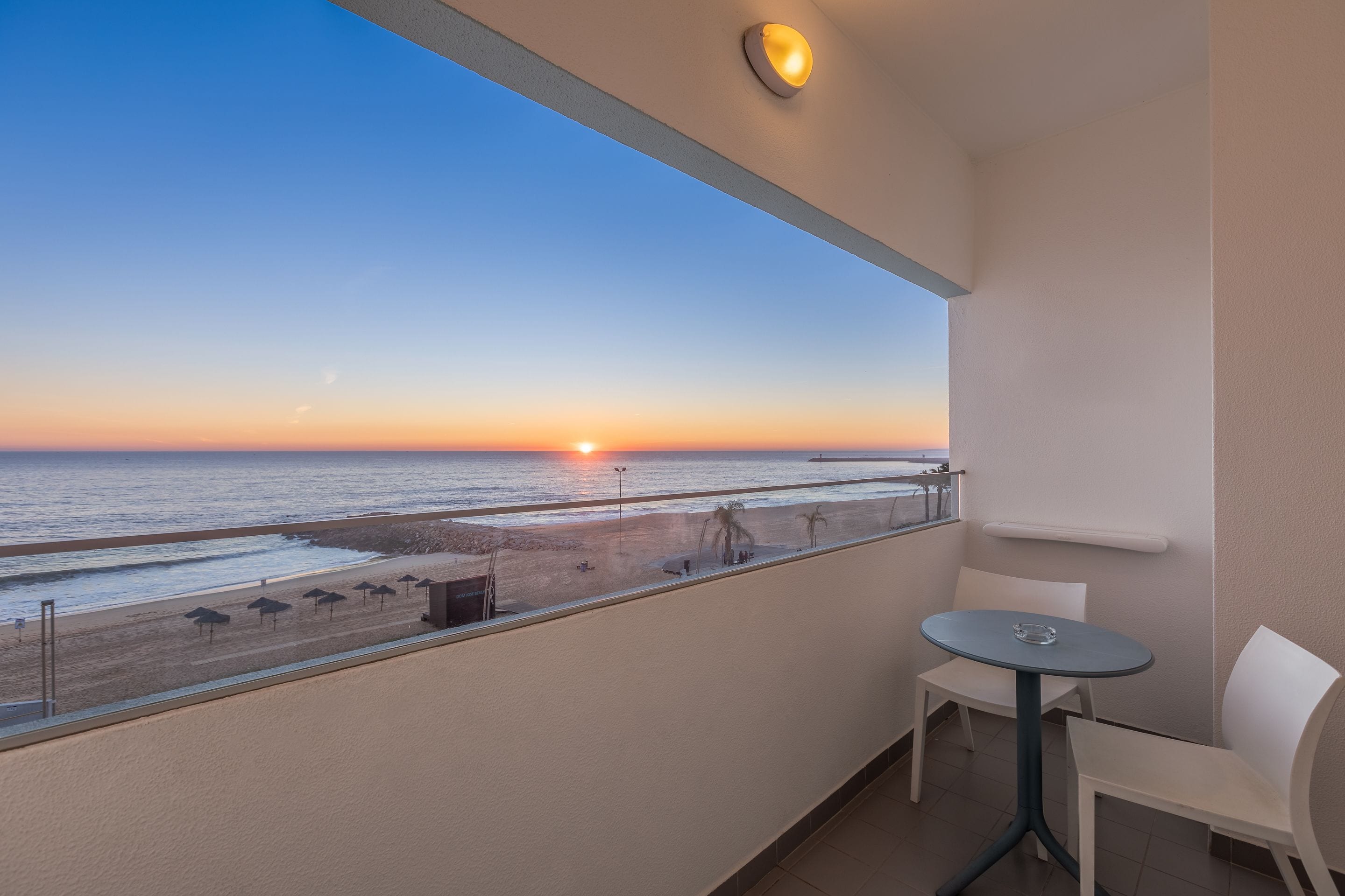 A balcony with a view of the sunset over the beach, a table, and two chairs.