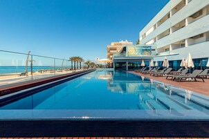 Ein Schwimmbecken mit Liegestühlen und Sonnenschirmen, ein Strand und ein Hotelgebäude mit Balkonen.