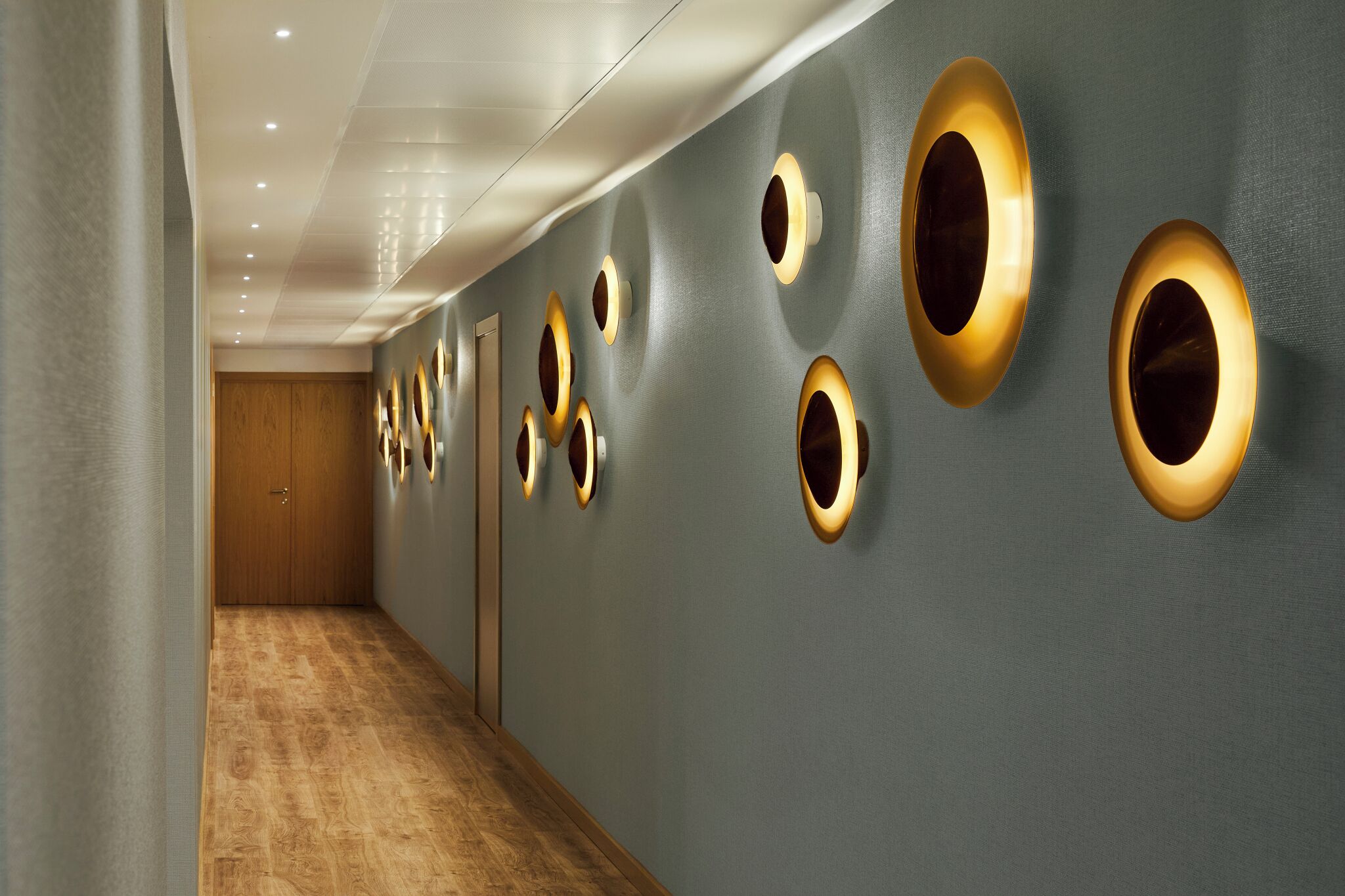 Un couloir doté d’un plancher en bois et d’un mur gris orné de luminaires circulaires.