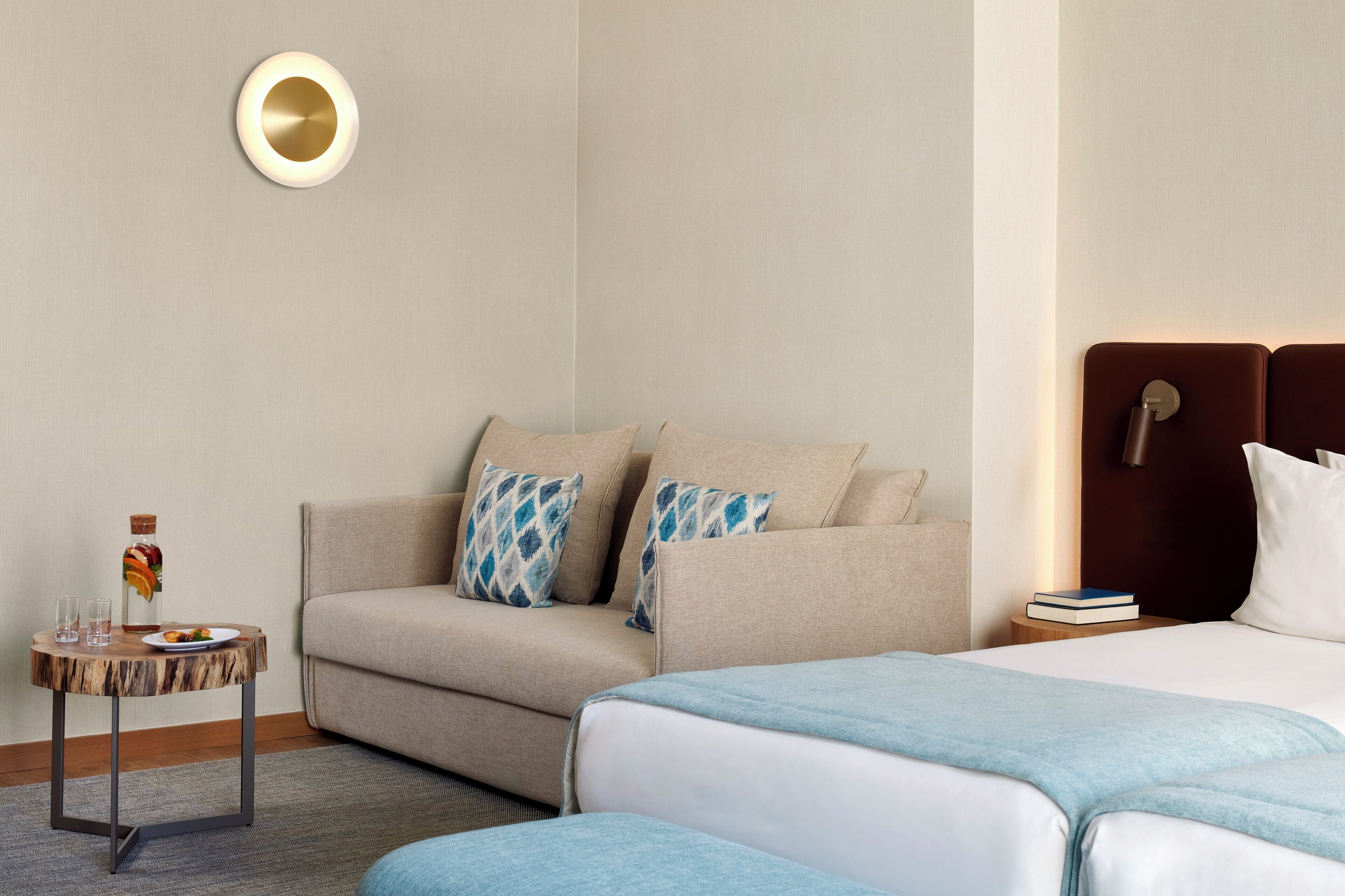 A hotel room with a sofa, a small round table with a bottle and glasses, a bed with a headboard, and a wall-mounted light fixture.