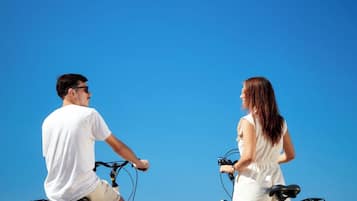 Deux personnes à vélo, se faisant face sur une terrasse en bois, avec un ciel bleu dégagé et l’océan en arrière-plan.