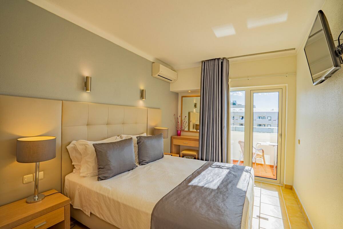 Habitación de hotel con una cama grande, lámparas de noche, un televisor de pantalla plana, un equipo de aire acondicionado, una puerta corrediza de vidrio a un balcón y una pequeña mesa con una silla.
