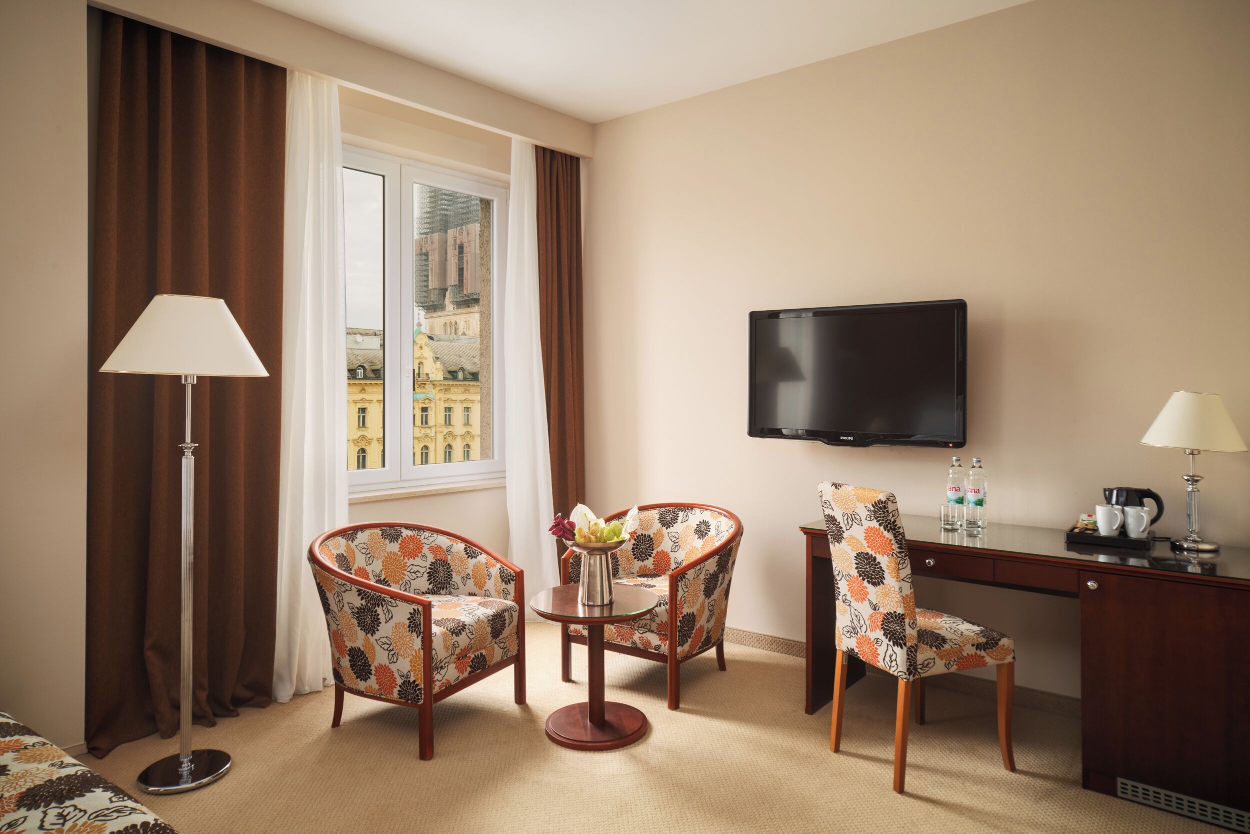 A hotel room with a desk, two armchairs, a TV, and a window with curtains.