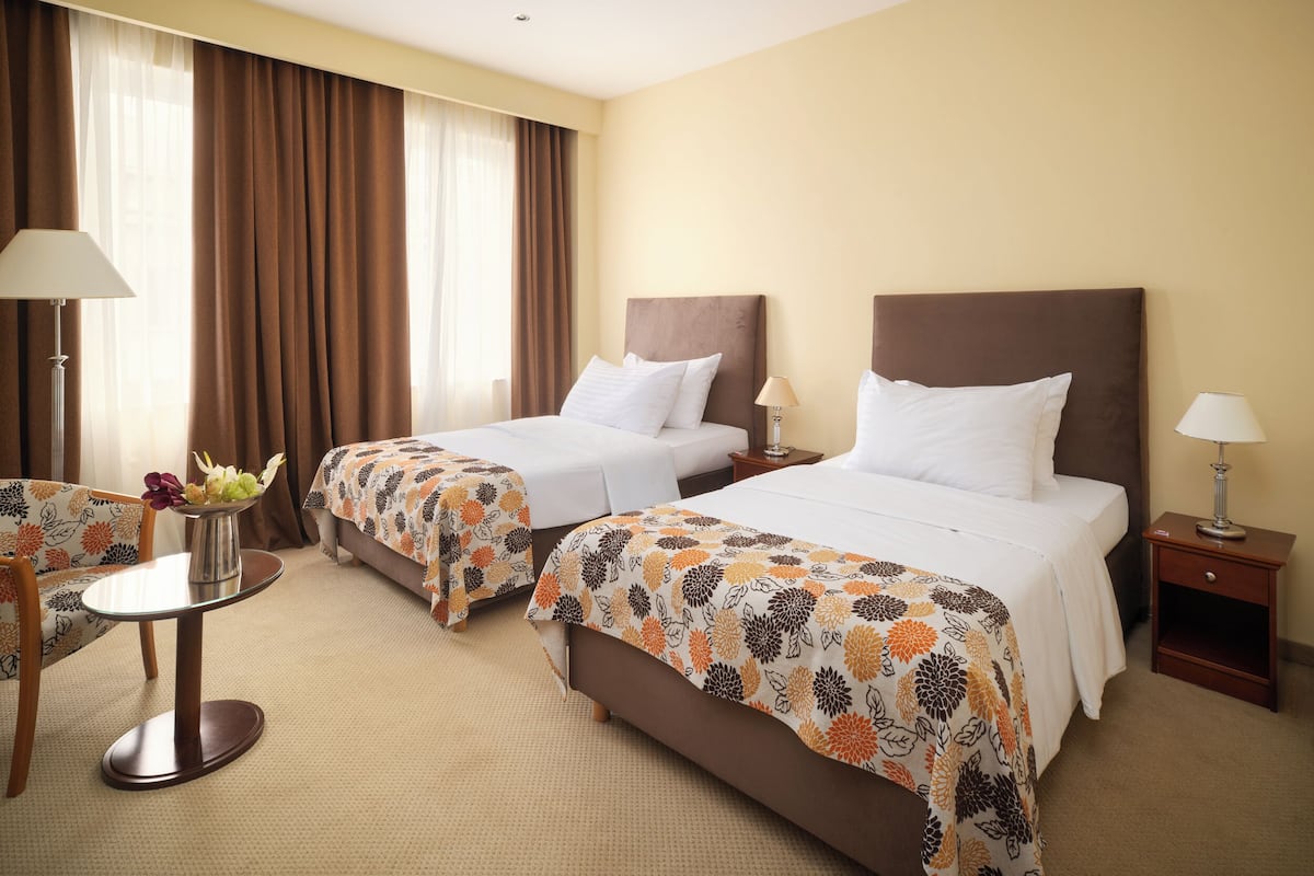 A hotel room with two beds, a small round table, a chair, and a nightstand with a lamp.