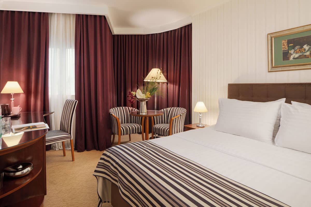 A hotel room with a large bed, two armchairs, a small table with a lamp, and a desk with a lamp.