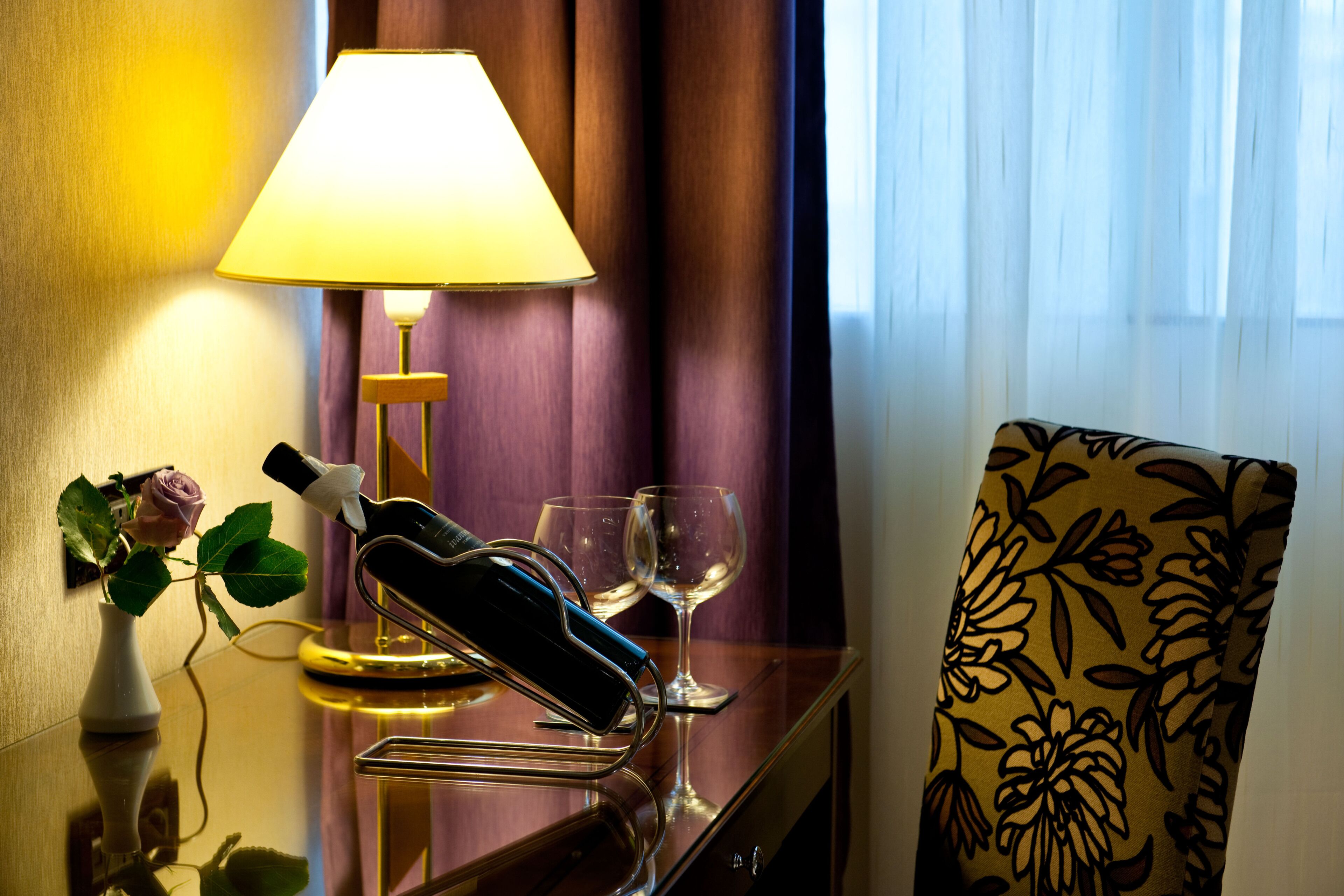Habitación de hotel con una mesita de noche, una lámpara, un florero con una rosa, dos copas de vino y una silla con estampado floral.