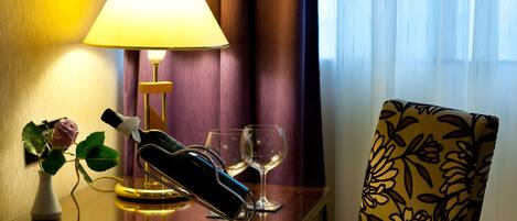 A hotel room with a bedside table, a lamp, a vase with a rose, two wine glasses, and a floral-patterned chair.
