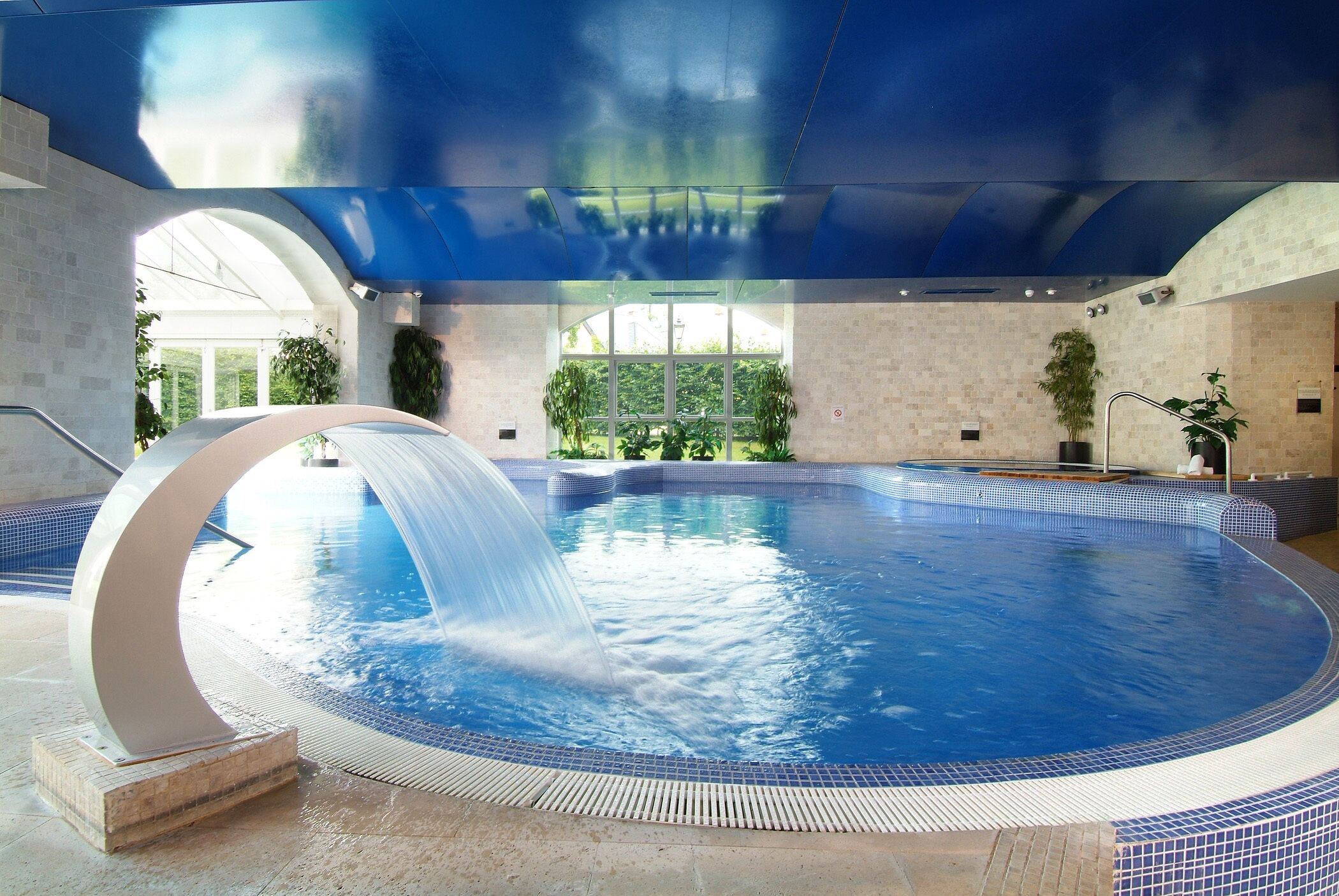 Una grande piscina interna con una cascata, circondata da pareti piastrellate e piante in vaso.