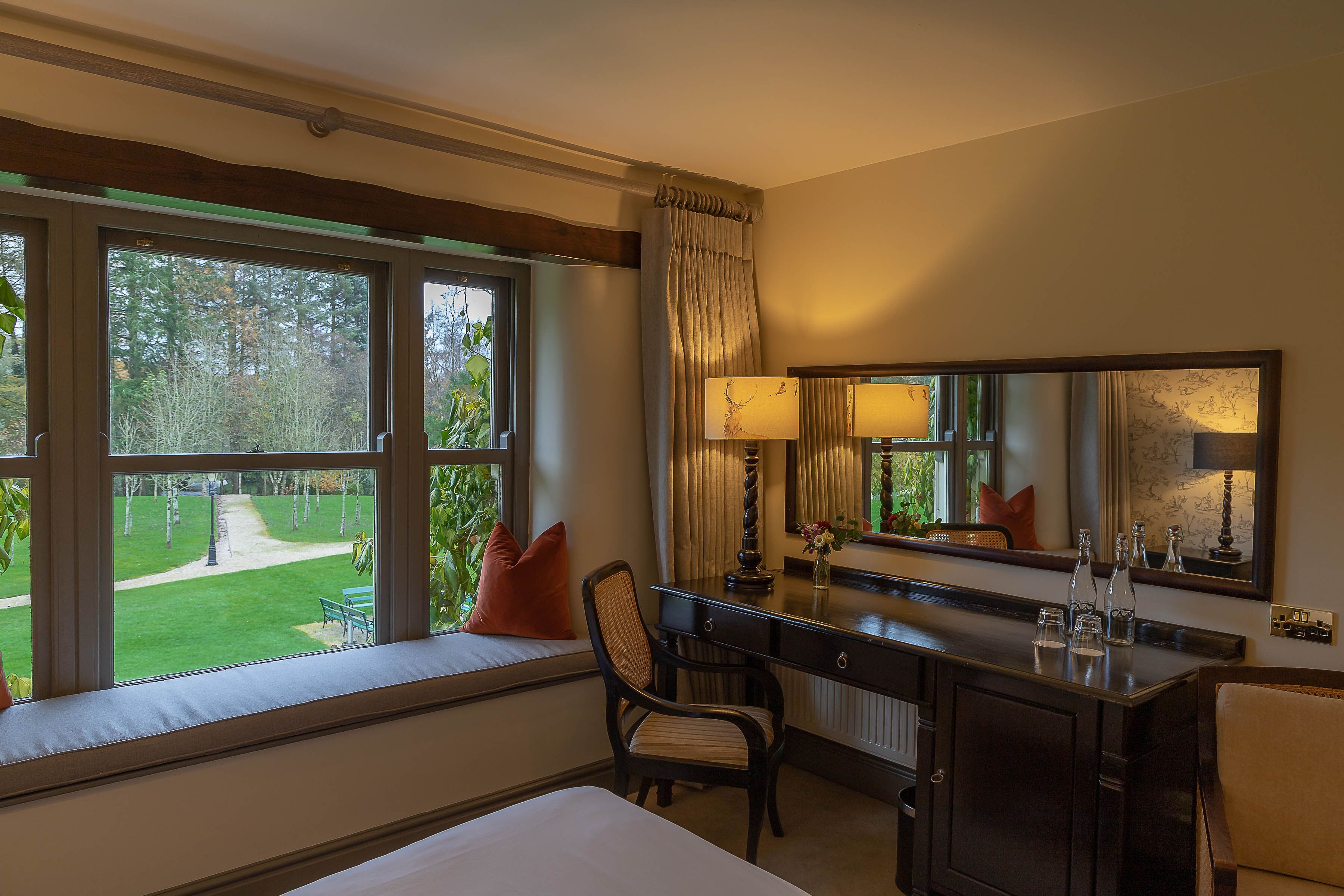 A hotel room with a large window, a desk, a chair, a bed, and a mirror.
