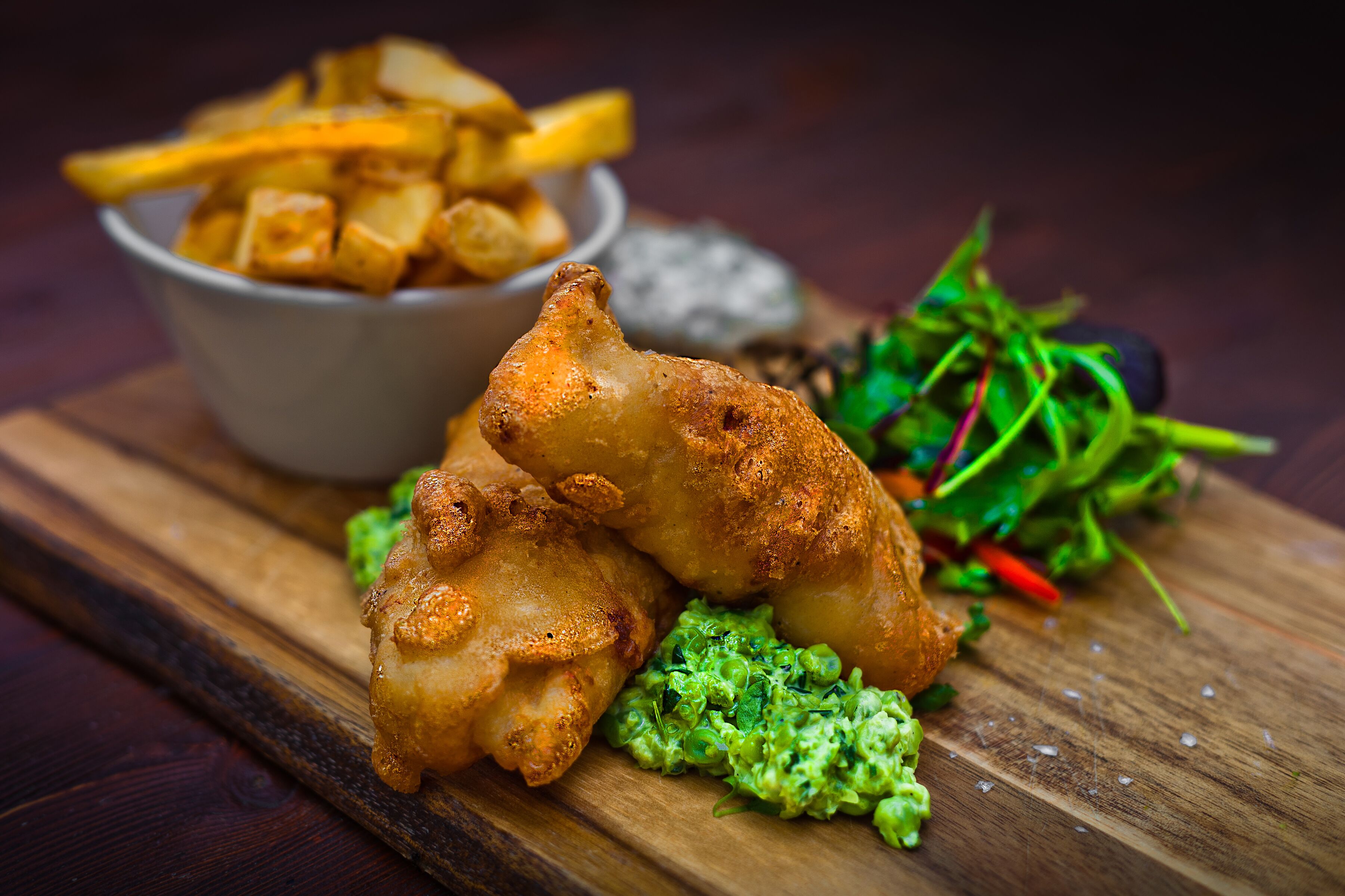 Una tabla de madera con comida frita, incluyendo un trozo de pollo frito y papas fritas, junto a una ensalada.