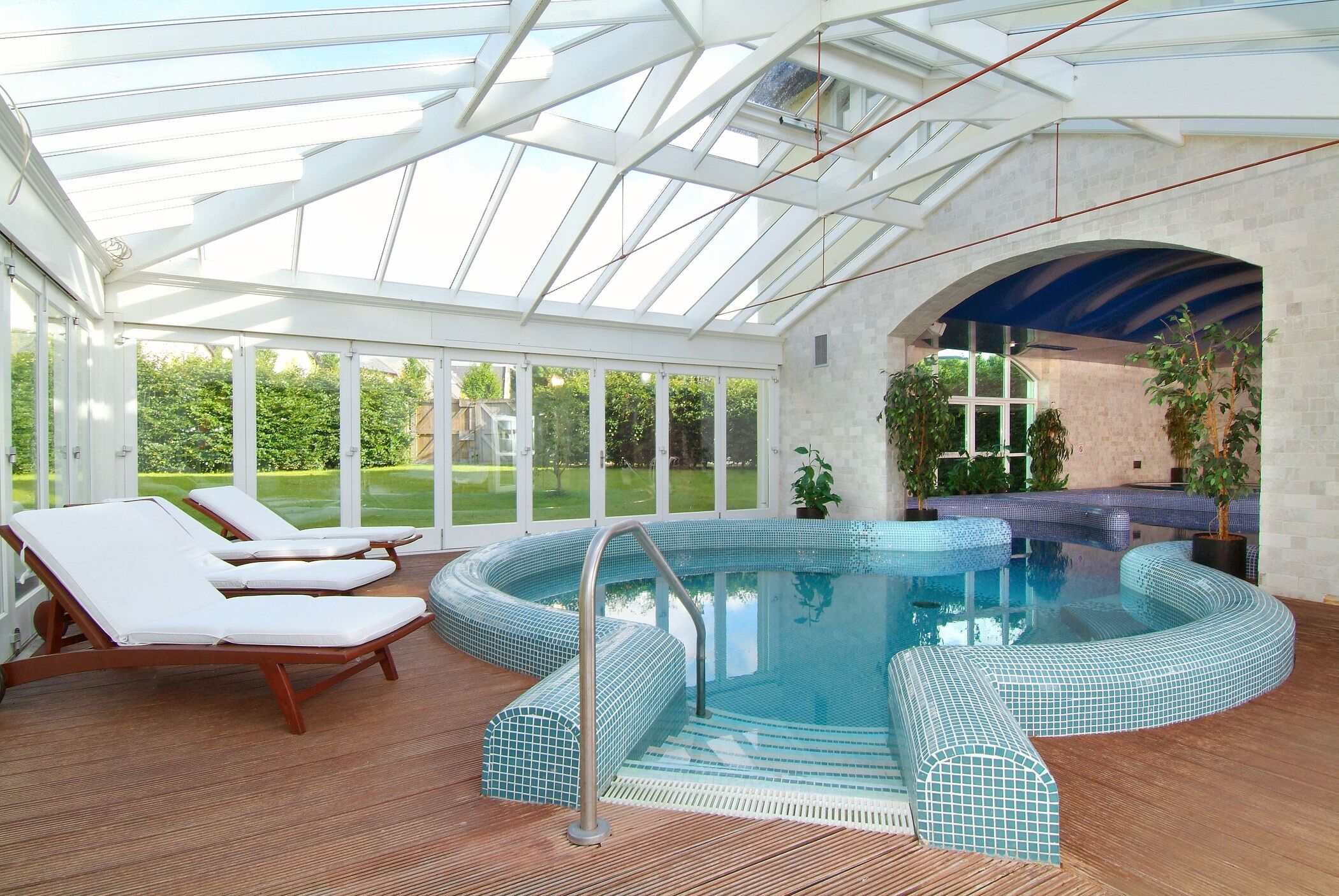 A sunlit indoor swimming pool with lounge chairs and a glass ceiling.