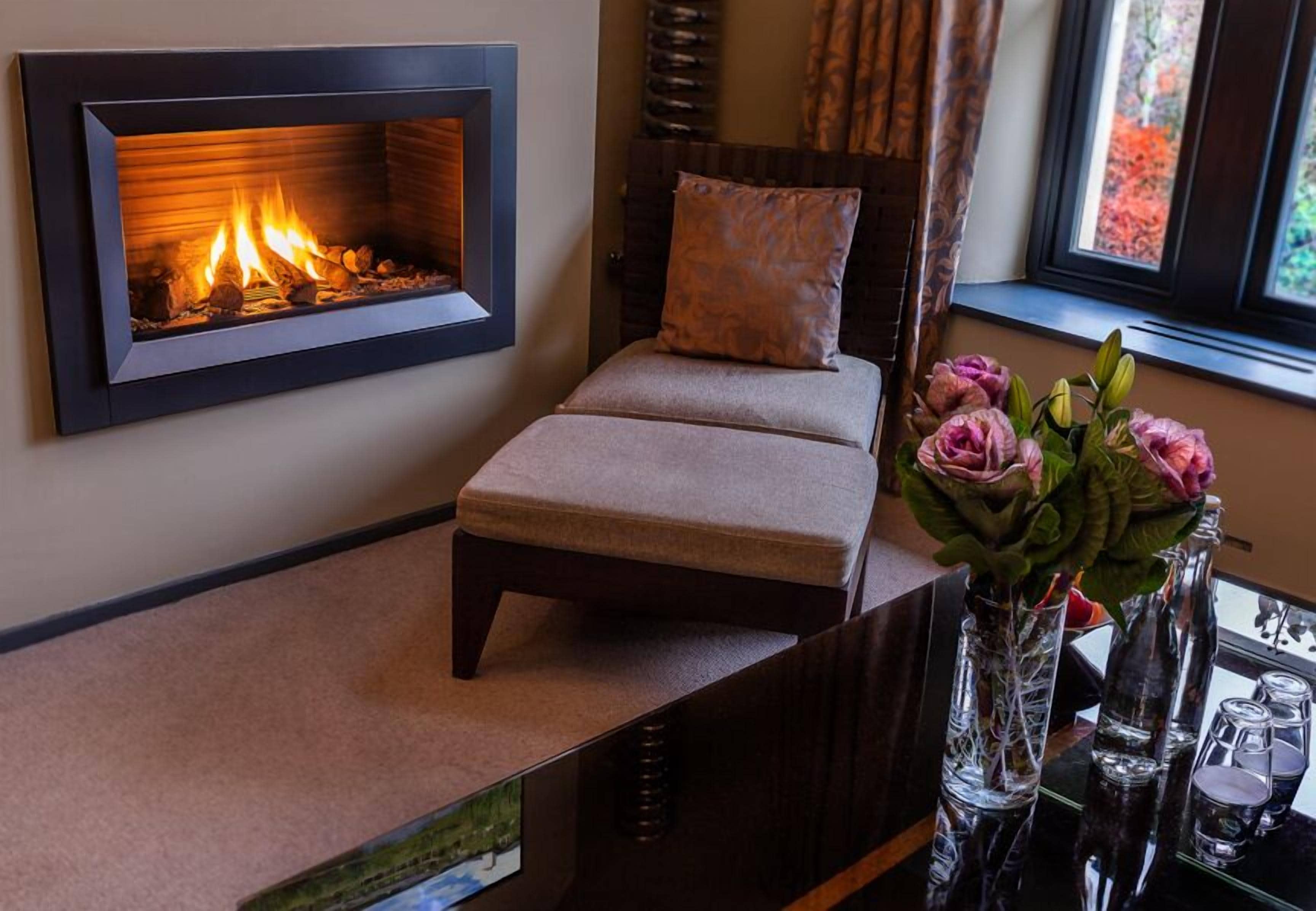 Una habitación con chimenea empotrada, un banco acolchado, un florero con flores y una ventana con follaje otoñal.