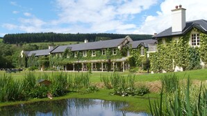 A large, ivy-covered building with a pond in front, surrounded by lush greenery and a forested hill in the background. - Brooklodge & Macreddin Village (Macreddin Village)