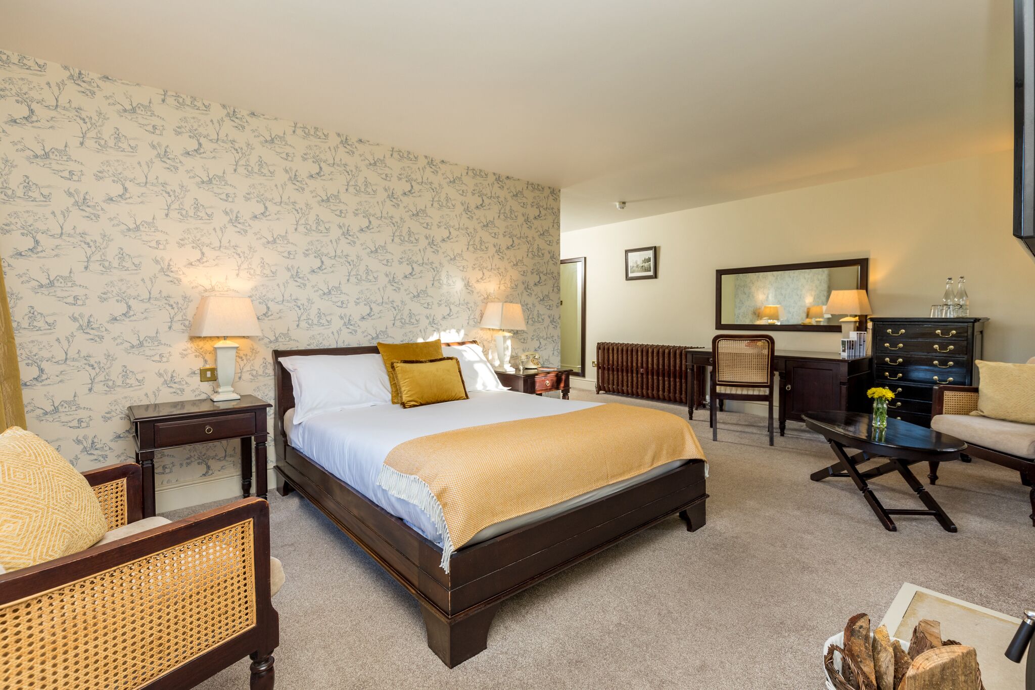Quarto de hotel com uma cama grande, duas mesas de cabeceira com abajures, uma cômoda de madeira, uma cadeira e uma pequena mesa com um vaso.