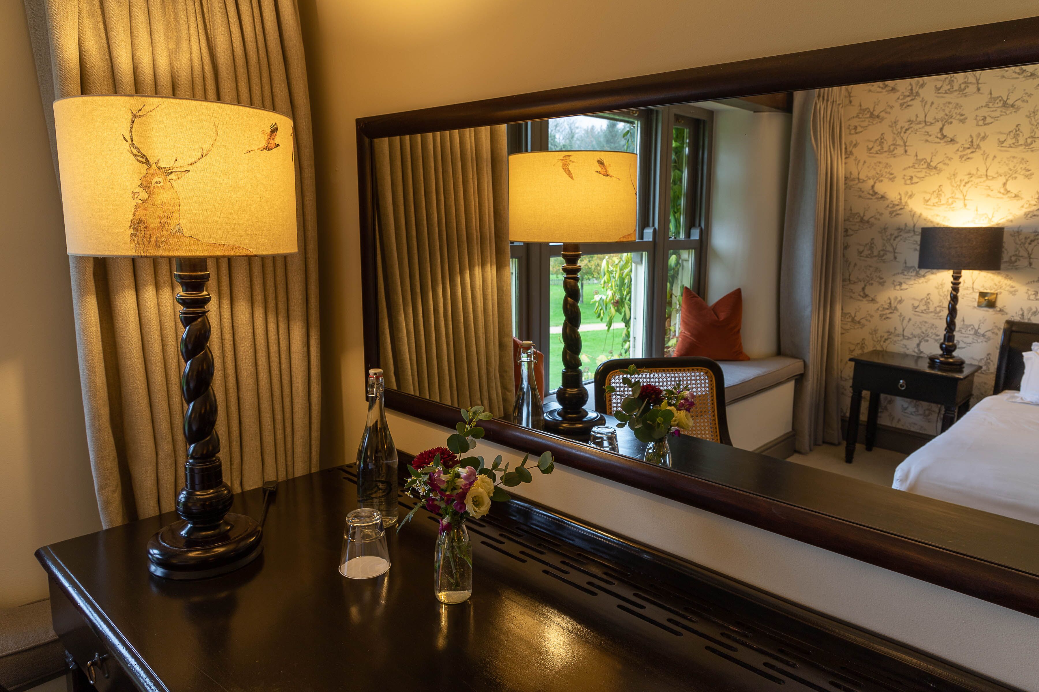 A hotel room with a large mirror reflecting the room's decor, a wooden dresser with a lamp, a vase with flowers, and a bed with a pillow.