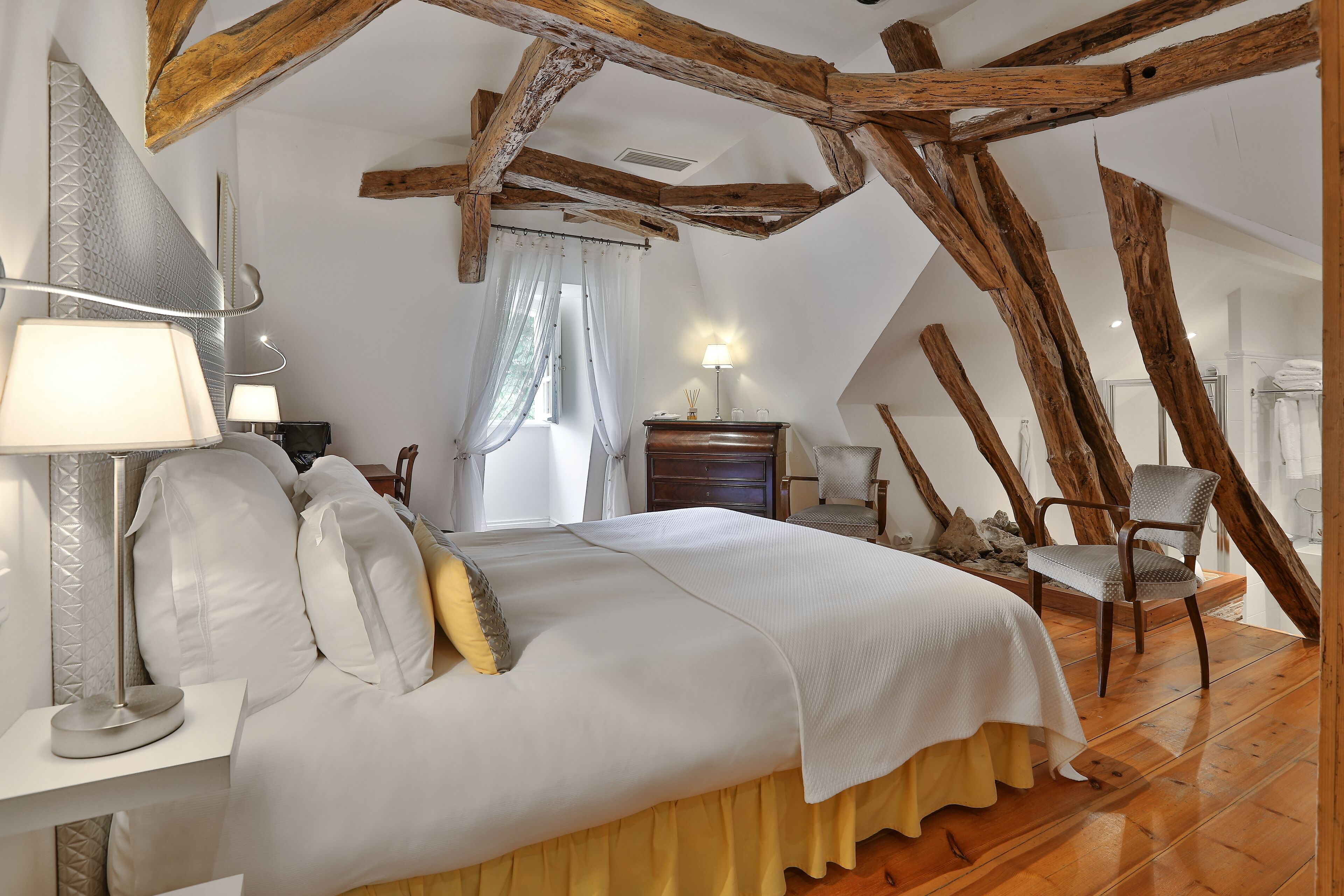 A bedroom with a wooden ceiling, a bed with white bedding, a nightstand with a lamp, and a chair.