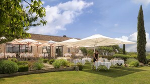 An outdoor dining area with tables and umbrellas, surrounded by greenery and stone buildings. - La Chartreuse du Bignac (Saint-Nexans)