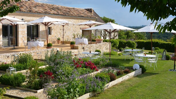 A stone building with outdoor seating and umbrellas, surrounded by a well-maintained garden with various plants and flowers. - La Chartreuse du Bignac (Saint-Nexans)