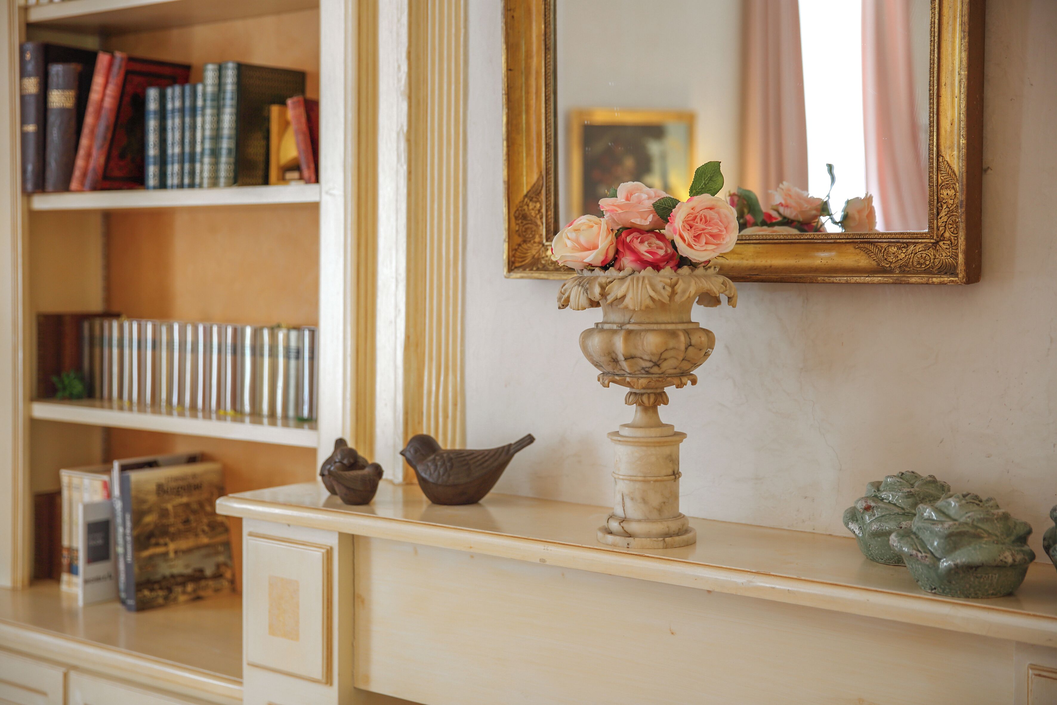 A bookshelf with books and decorative items, a mirror with a floral arrangement, and a small bird figurine on a dresser.