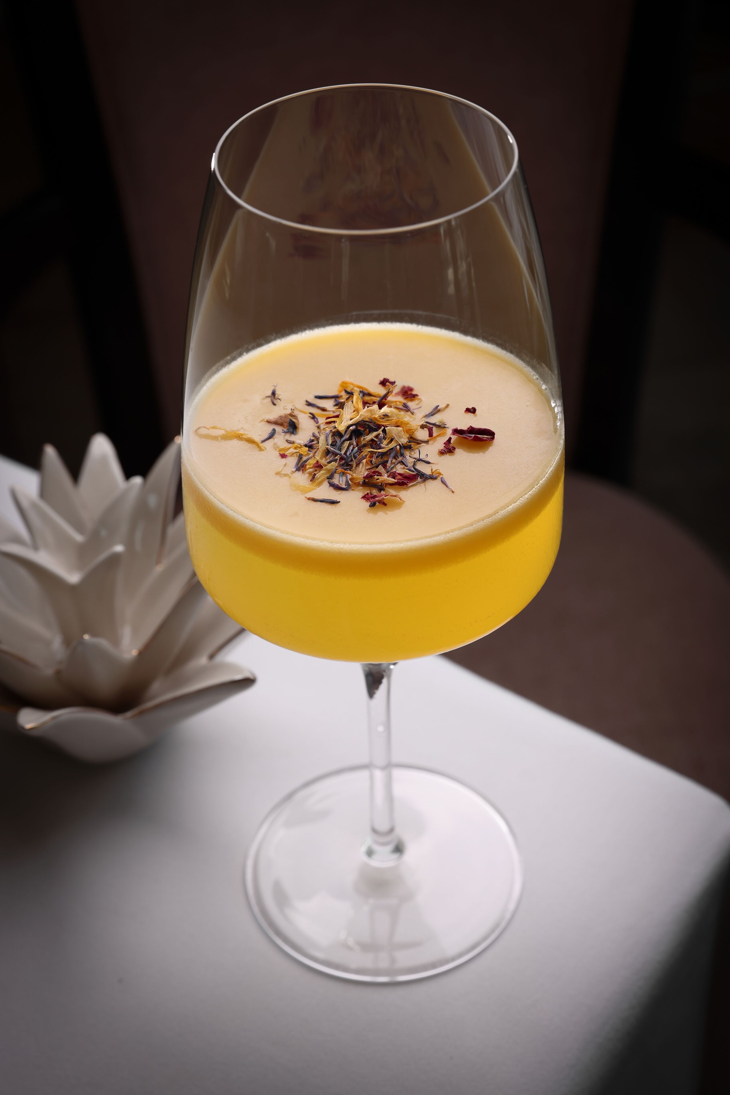 A cocktail in a stemmed glass with a yellow liquid and a garnish, placed on a white surface with a decorative object in the background.