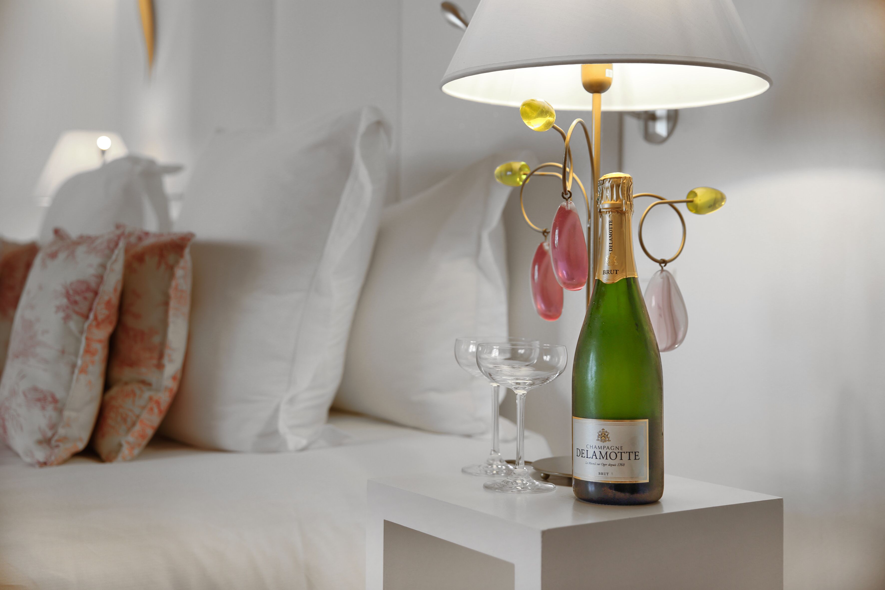 A bottle of champagne on a white table with a glass and a decorative lamp.