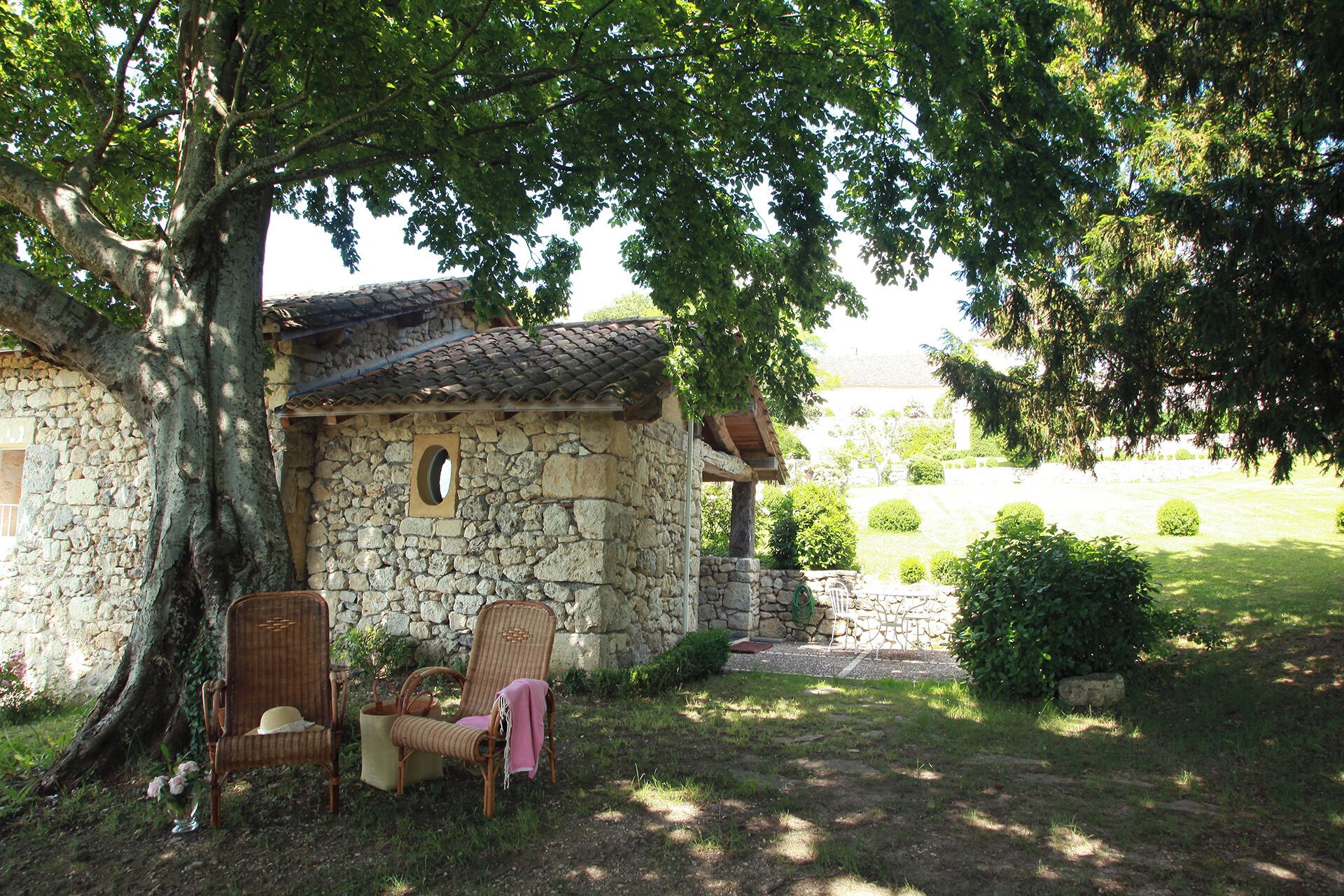 Un bâtiment en pierre, surmonté d’un toit en tuiles, entouré de verdure et agrémenté de quelques chaises en osier.