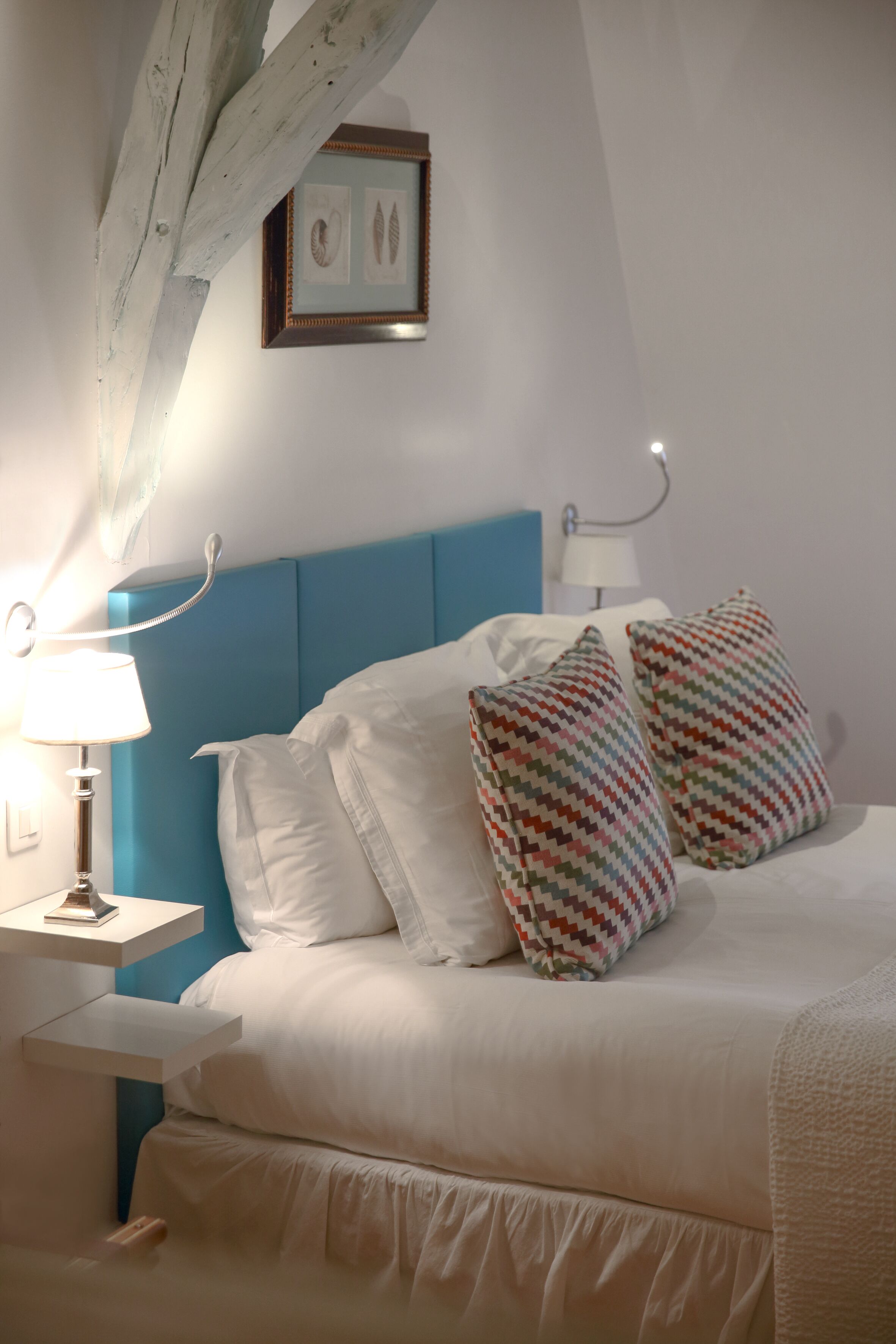 Un dormitorio con cama, ropa de cama blanca, cabecera azul, mesita de noche con lámpara y un cuadro en la pared.