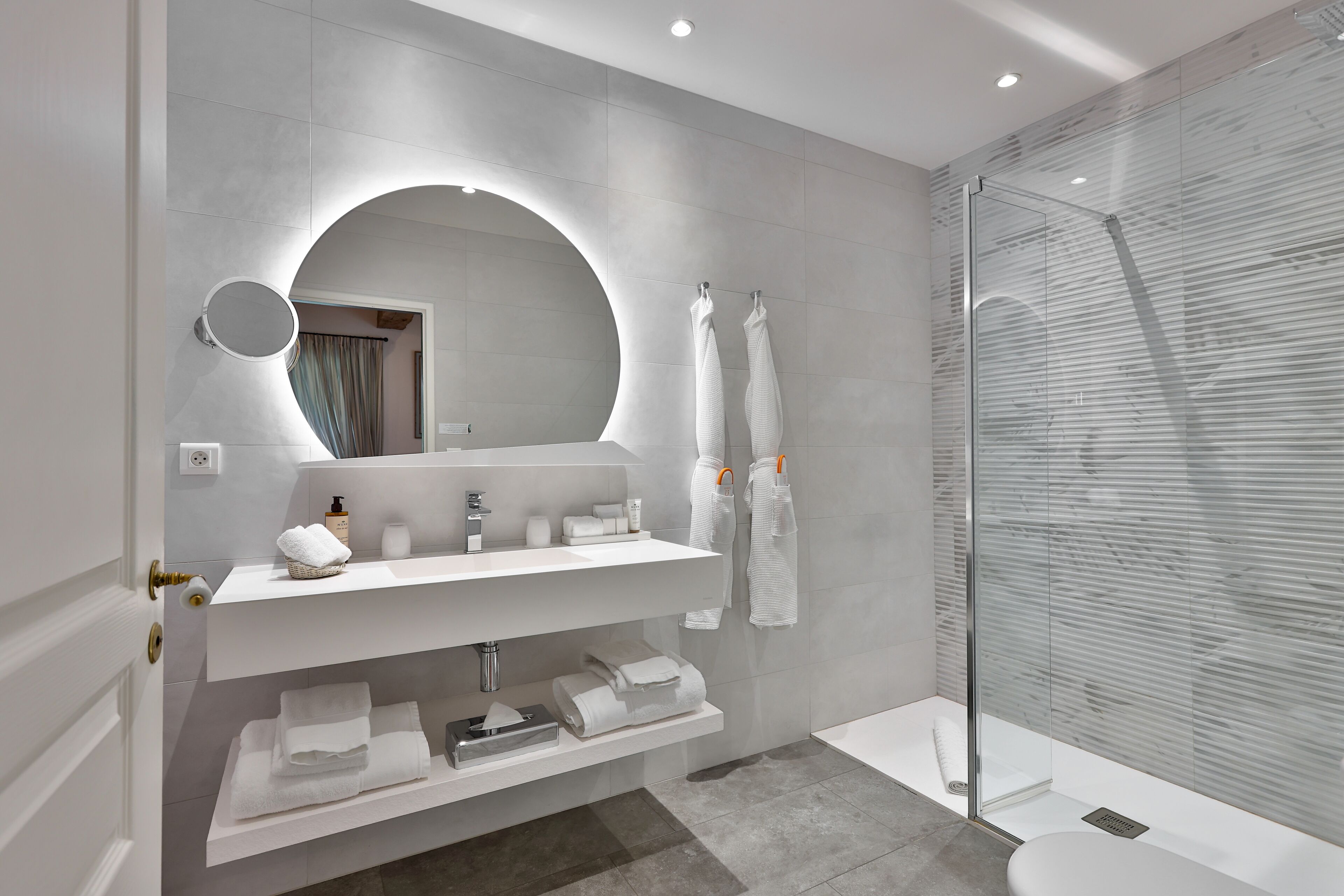 Un baño moderno con un espejo redondo, tocador blanco, toalleros y cabina de ducha con mampara de vidrio.