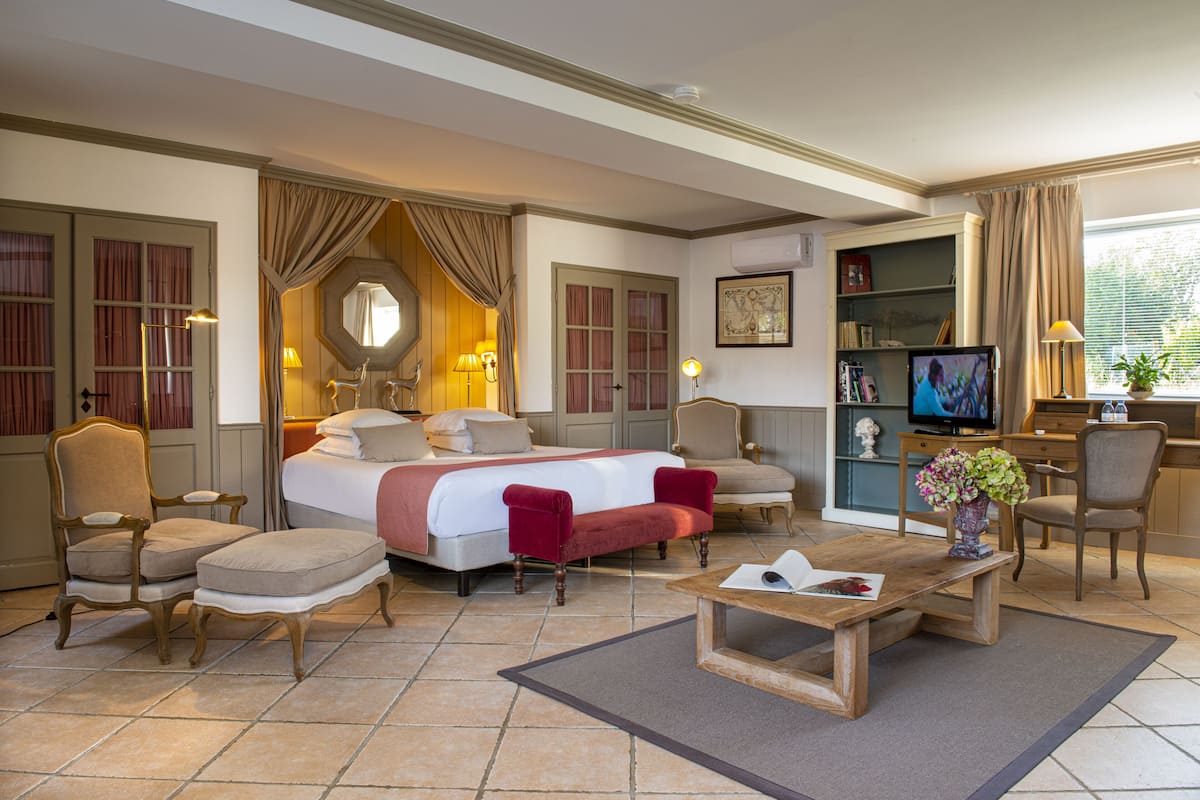 Ein Hotelzimmer mit einem großen Bett, zwei Sesseln, einem Holztisch, einem Fernseher und einem Bücherregal.