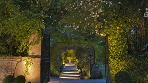 Un chemin qui traverse un jardin, avec un mur de pierre d’un côté et une grille métallique de l’autre.