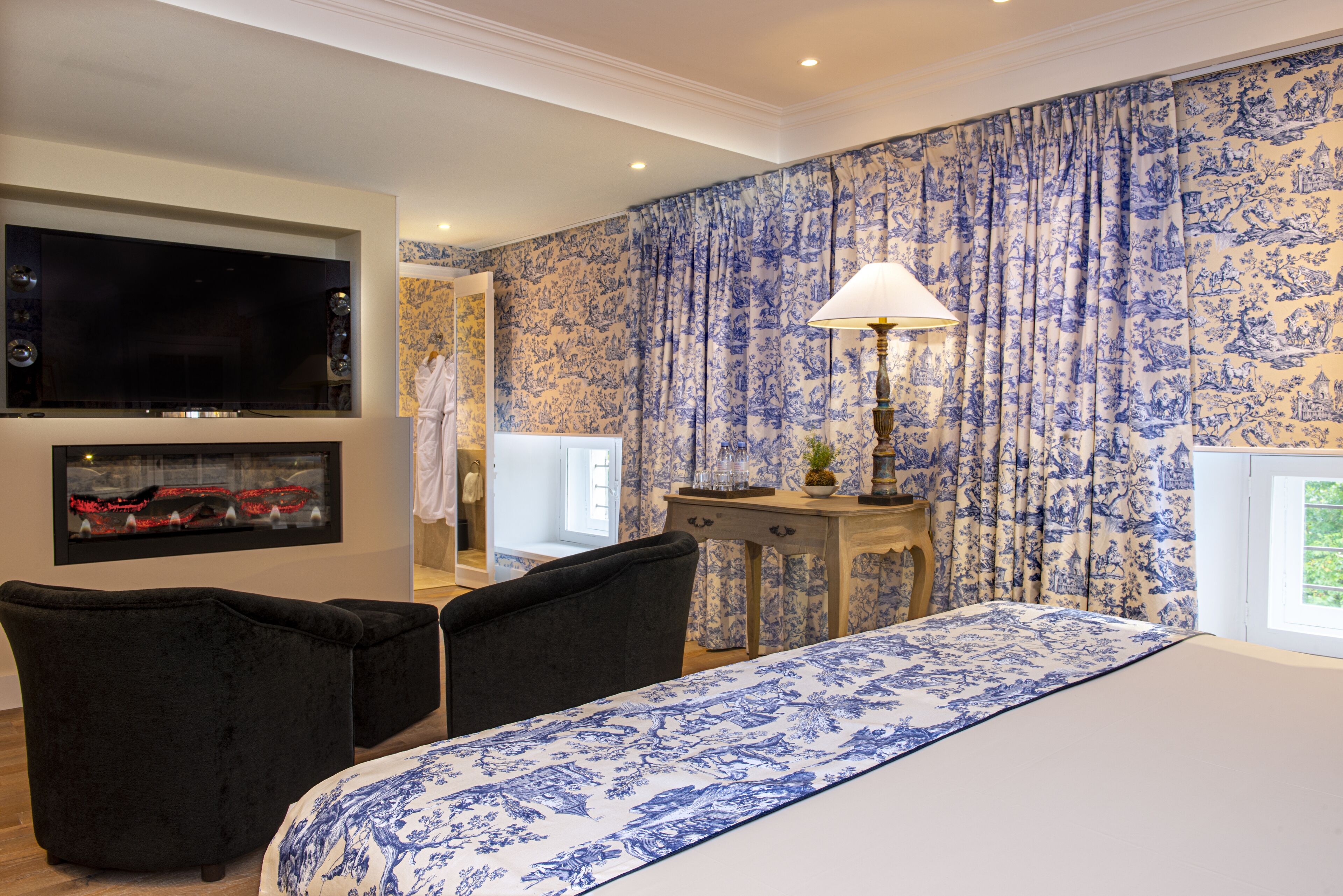 A bedroom with a large bed, two black chairs, a TV mounted on the wall, and a desk with a lamp.