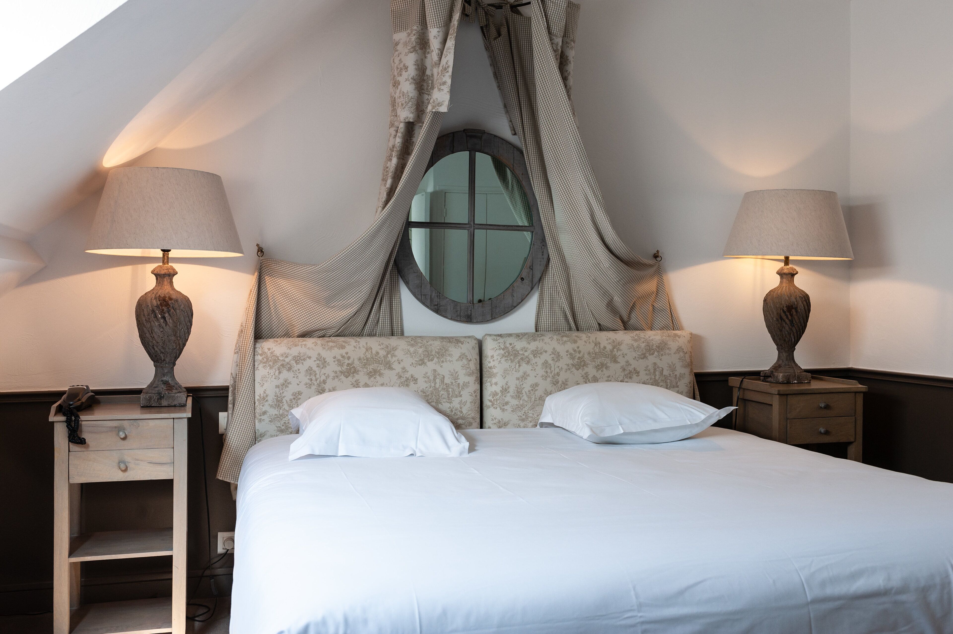 Una cama bien hecha con ropa blanca, dos lámparas de noche, una mesita de noche con una lámpara y una ventana con cortina.