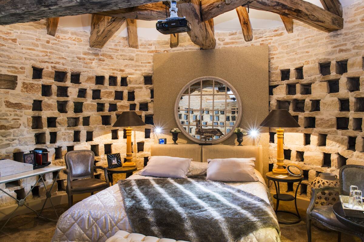 Ein gemütlicher Raum mit Steinwand, einem runden Spiegel, einem Bett mit Felldecke, zwei Stühlen, einem kleinen Tisch und einer Lampe.