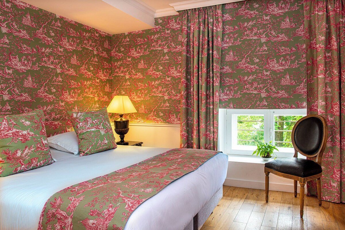 Een slaapkamer met bloemenbehang, een bed met een wit- en groen beddegoed, een houten stoel en een raam met uitzicht op groen.