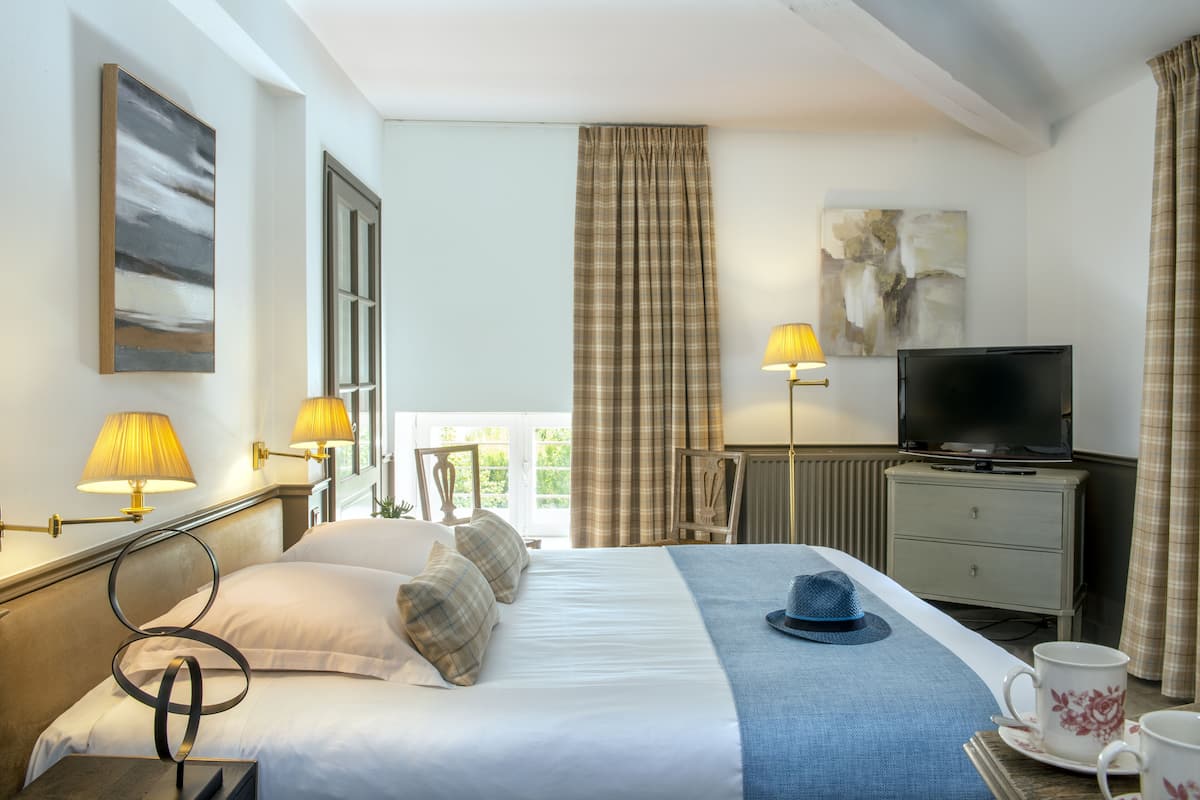 Ein Hotelzimmer mit Bett, Fernseher, Schreibtisch mit Lampe und Fenster mit Vorhängen.