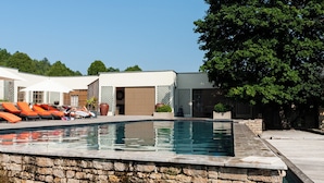 Une piscine aux murs en pierre, avec des chaises longues orange et un grand arbre en arrière-plan.