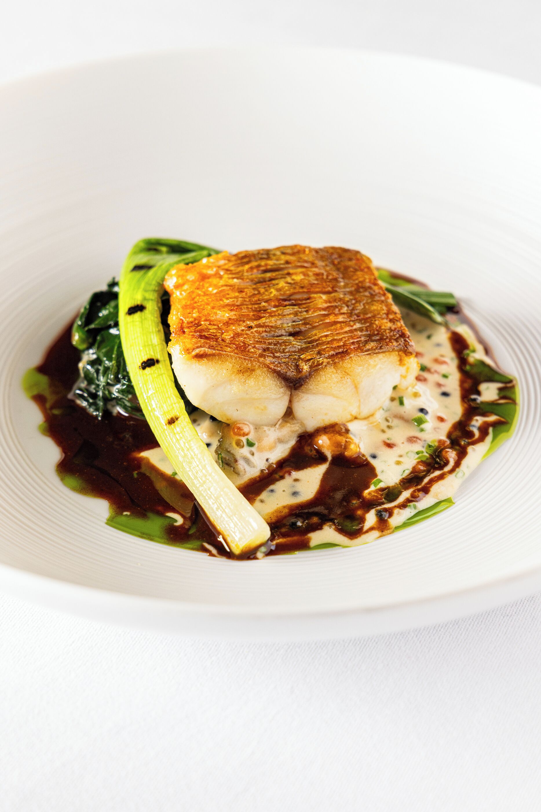 A white plate with a piece of fish, green vegetables, and a sauce.