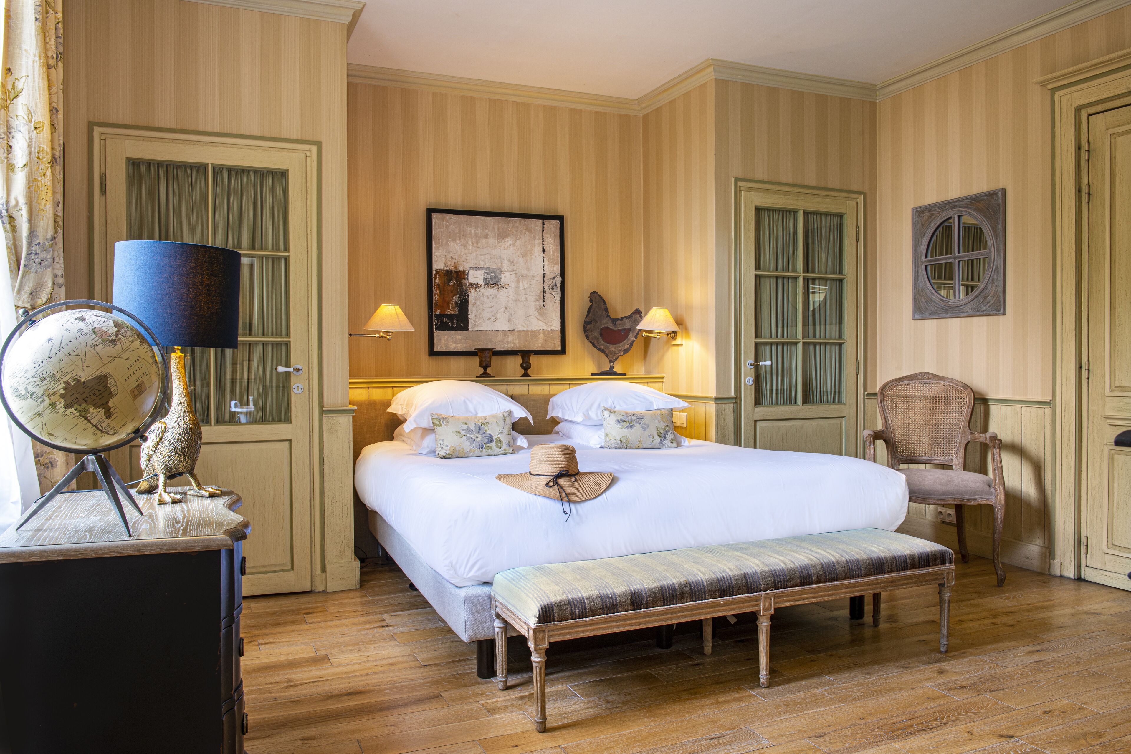 A neatly arranged bedroom with a large bed, bedside lamps, a globe, and a wicker chair.