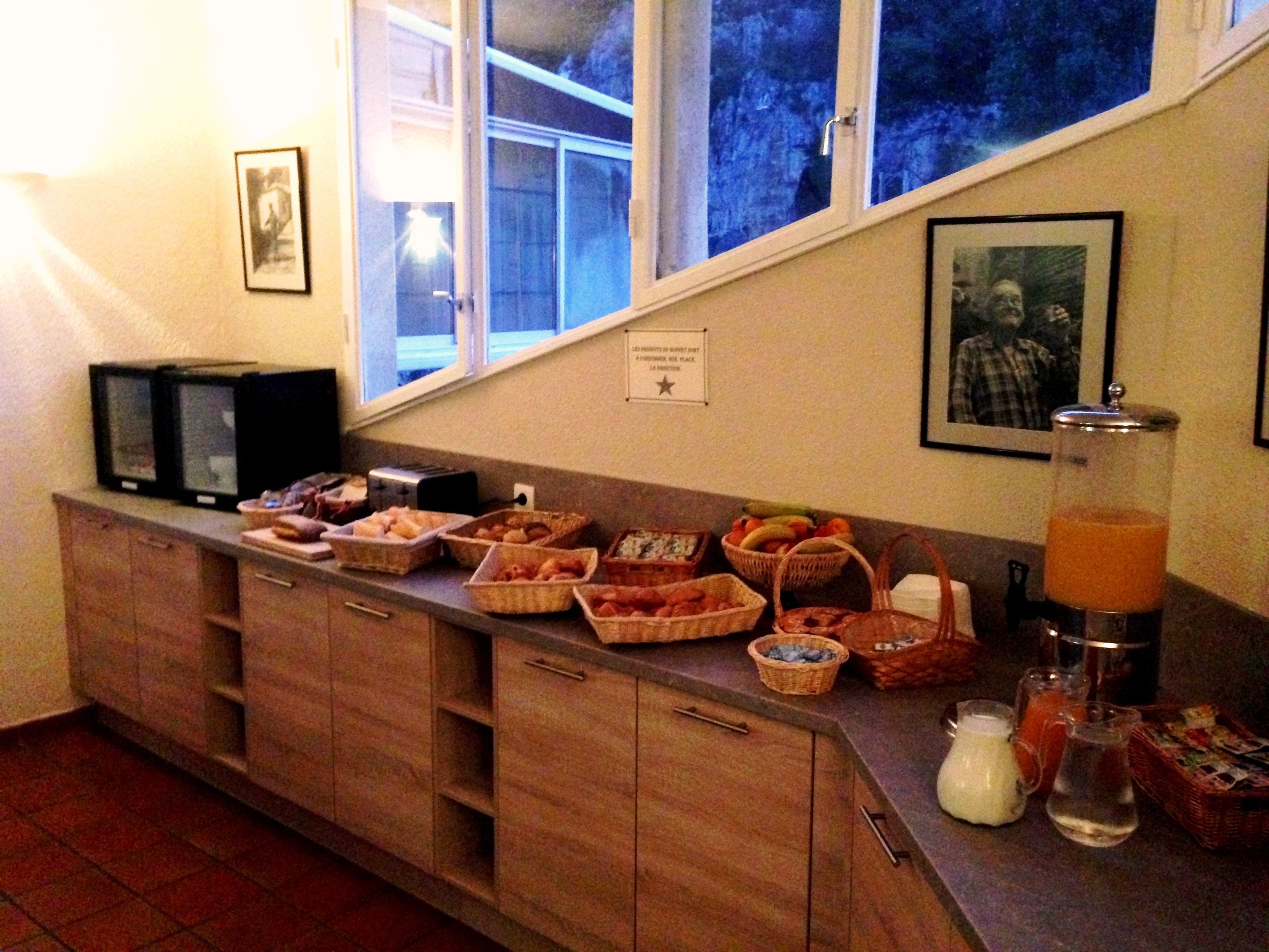 A kitchen with wooden cabinets, a countertop with various food items, a window with a view of trees, and a wall-mounted picture.